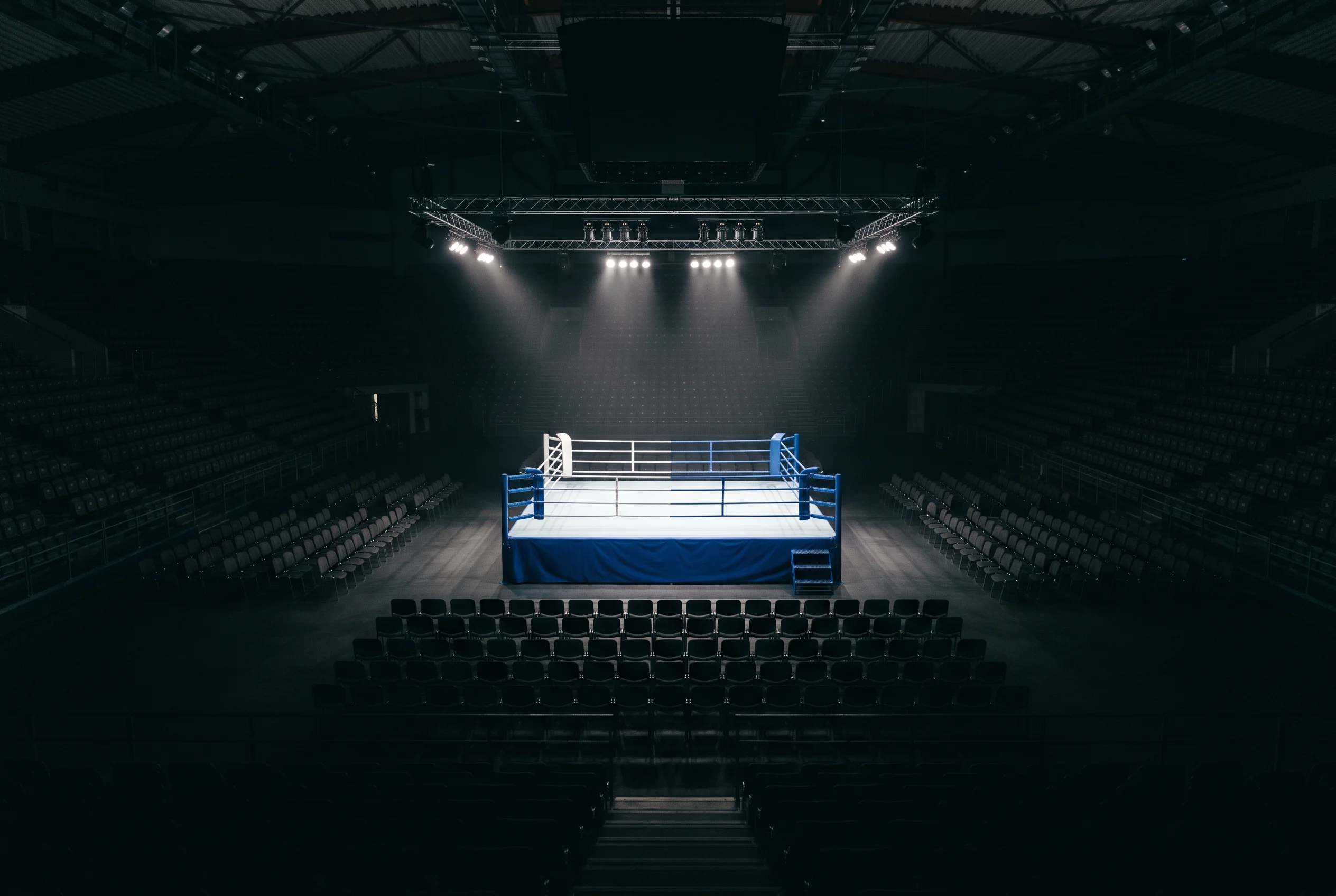Gran ring de boxeo iluminado en el centro de un pabellón con butacas vacías antes del evento